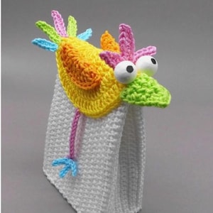 May include: A whimsical, handmade crochet towel topper in the shape of a colorful bird. The bird features a yellow, orange, and pink head with large googly eyes and a green beak. The towel is white and has a textured pattern.