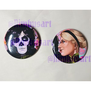 May include: Two circular pin-back badges featuring illustrated characters. The left badge shows a figure with dark hair and skull-like face paint. The right badge depicts a profile view of a blonde person with a black headband, both on pink backgrounds.