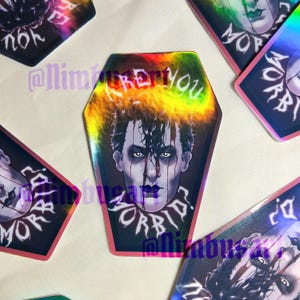 May include: A coffin-shaped holographic sticker featuring a stylized illustration of a person with dark hair, pale skin, and dark makeup. The text "ARE YOU MORBID?" is printed in a white gothic font above and below the portrait.