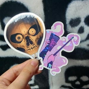 May include: Two die-cut stickers featuring a hot air balloon shaped like a human skull with dark eye sockets and a second sticker showing a hand holding a purple book with the word Slut printed on the cover and a wand emitting pink smoke.