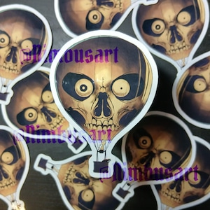 May include: A collection of die-cut stickers featuring a hot air balloon shaped like a human skull. The balloon has a beige and brown gradient, dark eye sockets, and a small black basket underneath. The text "Nimousart" is printed in purple.