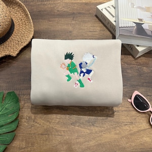 May include: A beige sweatshirt with an embroidered design featuring two cartoon characters in green and blue outfits. The characters are in action poses, with one holding an orange ball. The sweatshirt is folded on a wooden surface, with a straw hat and sunglasses nearby.