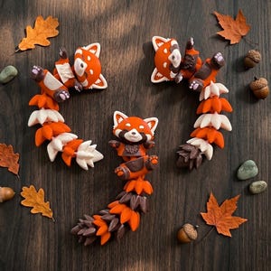 Peut inclure: Trois figurines de pandas roux aux détails orange, blancs et bruns sont disposées sur une surface en bois foncé, entourées de feuilles d'automne, de glands et de petites pierres. Les figurines ont un design ludique et cartoon.