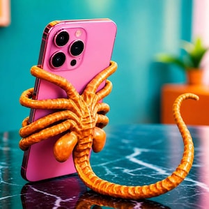 Könnte beinhalten: Ein pinkfarbenes Smartphone wird von einem orangefarbenen, tentakelartigen Handyhalter gehalten. Das Telefon hat eine Dreifach-Kamera. Der Halter ist so gestaltet, dass er einem Facehugger aus der Alien-Filmreihe ähnelt. Das Telefon und der Halter stehen auf einer dunkelblauen Marmoroberfläche.