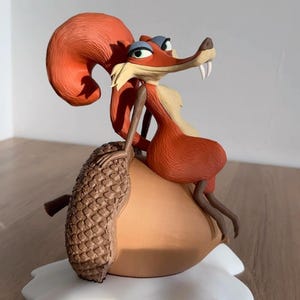 Op de afbeelding: Gedetailleerd beeldje van een cartoon eekhoorn karakter, met roodachtig-oranje vacht en een lange, gebogen staart. De eekhoorn zit op een grote eikel, met een dennenappel in zijn greep. De basis is wit en heeft de vorm van een wolk.