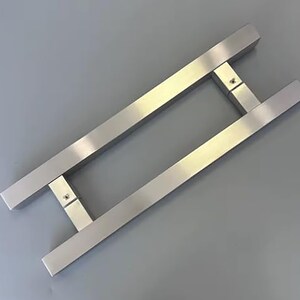 May include: A brushed stainless steel door pull handle. The handle has a rectangular bar design with a square connecting piece and is mounted on a gray surface. The handle is designed for a modern aesthetic.