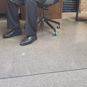 K&ouml;nnte beinhalten: Eine transparente, rechteckige B&uuml;rostuhlmatte auf einem Teppichboden. Die Matte befindet sich unter einer Person, die in einem schwarzen B&uuml;rostuhl sitzt. Die Person tr&auml;gt eine schwarze Hose und schwarze Schuhe.