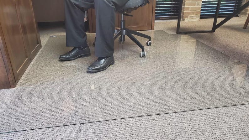 K&ouml;nnte beinhalten: Eine transparente, rechteckige B&uuml;rostuhlmatte auf einem Teppichboden. Die Matte befindet sich unter einer Person, die in einem schwarzen B&uuml;rostuhl sitzt. Die Person tr&auml;gt eine schwarze Hose und schwarze Schuhe.
