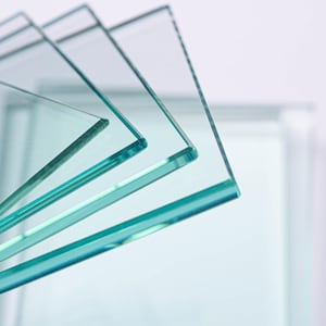 May include: A stack of clear glass squares, each with a slightly green tint, are arranged in a fan shape. The edges of the glass are visible, showcasing their thickness and smooth texture. The background is a soft, blurred white.