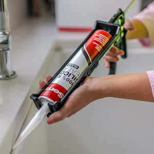 May include: A black caulking gun holding a red and white tube of 100% silicone sealant. The sealant tube is in a black caulking gun, ready for use. The image shows a close-up of the sealant being applied to a white surface.