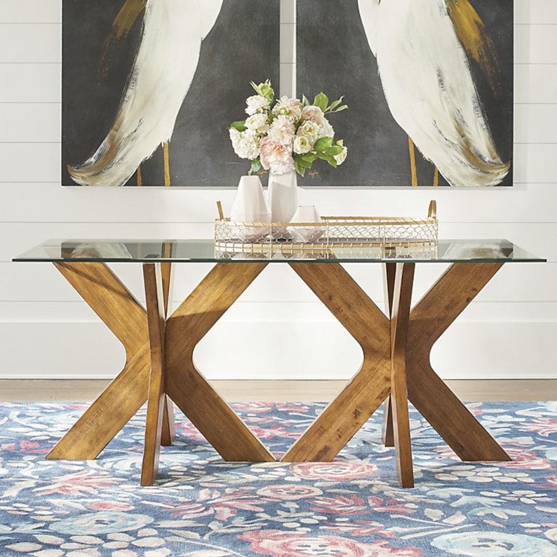K&ouml;nnte beinhalten: Ein rechteckiger Tisch mit Glasplatte und Holzgestell. Das Gestell hat ein X-f&ouml;rmiges Design in einem warmen Braunton. Der Tisch steht auf einem blauen Teppich mit Blumenmuster, mit Kunstwerken und Dekorationen im Hintergrund.