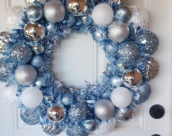 May include: A decorative wreath featuring silver, white, and blue baubles. The wreath is composed of various sizes of shiny and textured spheres, creating a sparkling festive decoration. It hangs on a white door.