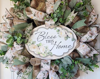 May include: A decorative wreath featuring a central oval sign with the text Bless This Home, surrounded by layers of mesh ribbon in gold and beige, floral patterned fabric ribbons, and artificial green eucalyptus foliage.