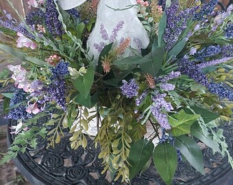 May include: A white rabbit ornament surrounded by purple, pink, and green artificial flowers and foliage. The arrangement is on a black, ornate metal table, creating a spring or Easter theme.