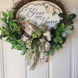 May include: A round grapevine wreath featuring a central oval sign with the text "Bless THIS Home" surrounded by white floral illustrations. The wreath is decorated with green faux leaves and a multi-layered bow with a floral and leaf pattern.