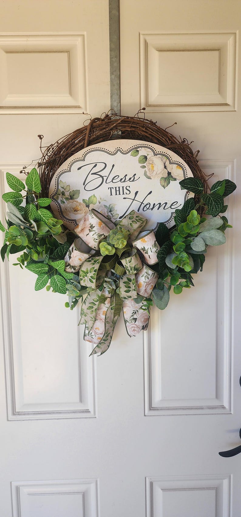 May include: A round grapevine wreath featuring a central oval sign with the text "Bless THIS Home" surrounded by white floral illustrations. The wreath is decorated with green faux leaves and a multi-layered bow with a floral and leaf pattern.