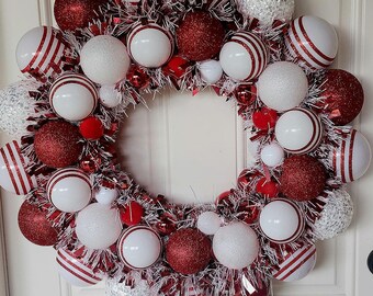 May include: A Christmas wreath with red, white, and silver baubles. The wreath is decorated with large, glittery red and white striped balls, small white and silver spheres, and red pom-poms, creating a festive holiday decoration.