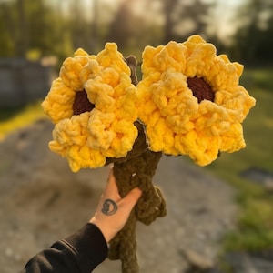 May include: Two handmade crochet sunflowers with vibrant yellow petals and brown centres. The stems are a dark green colour. The flowers are held in a person's hand against a blurred outdoor background.