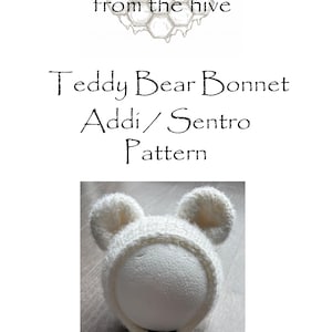 May include: A white knitted teddy bear bonnet with two rounded ears. The text "Teddy Bear Bonnet Addi/Sentro Pattern" is displayed above the bonnet. The image also includes the text "from the hive".