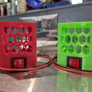 May include: Two electronic devices with red and green honeycomb-patterned housings. Each has a red on/off switch and a clear bulb visible through the hexagonal cutouts. Black and red wires extend from the base.