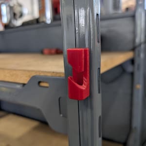 May include: A close-up of a red 3D-printed hook attached to a grey metal shelf support. The hook is designed to fit into the pre-drilled slots of the shelf. The background shows a wooden shelf and other items.