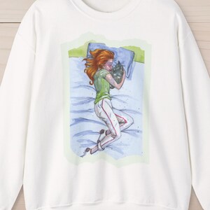 Può includere: Felpa bianca con un'illustrazione ad acquerello di una persona dai capelli rossi sdraiata su un letto, che abbraccia un gatto. La persona indossa un top verde e pantaloni bianchi. L'opera è incorniciata da un bordo verde chiaro.