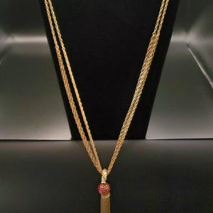 May include: Gold-toned necklace with a tassel pendant. The necklace features a braided chain design, with a red beaded accent at the top of the tassel. The necklace is displayed on a black mannequin.