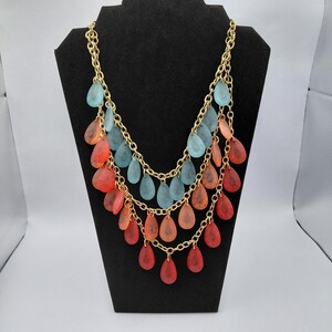 May include: A multi-strand necklace with a gold chain and teardrop-shaped pendants. The pendants are in shades of blue, orange, and red. The necklace is displayed on a black jewelry stand.
