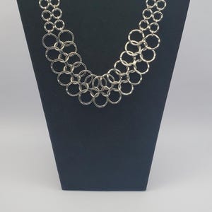 May include: Silver-toned necklace with a series of interconnected circles. The necklace is displayed on a black jewelry stand against a white background. The circles are of varying sizes, creating a layered effect.
