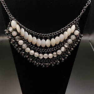 May include: A layered necklace with a gunmetal chain and off-white oval beads. The necklace is displayed on a black velvet bust, showcasing its multiple strands and intricate design. A stylish accessory.