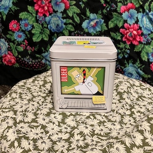 May include: A rectangular, light gray metal tin featuring a Dilbert cartoon on the front. The cartoon shows Dilbert inside a computer screen. The tin has a hinged lid and a keyboard design on the bottom. The text "REMEMBER TO LOWER EXPECTATIONS!" is also visible.