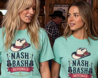 以下が含まれることがあります： カウボーイハット、2本のアコースティックギター、そして赤いバナーに「NASH BASH BRIDESMAID」の文字が描かれた、ライトティールカラーのTシャツ3枚。木製の壁がある素朴なバーで、3人がこのTシャツを着用しています。