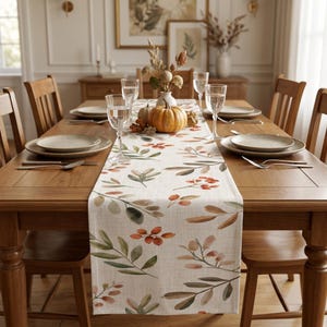 May include: A rectangular table runner featuring a printed pattern of watercolor-style autumn leaves and berries in shades of orange, brown, and green on a light beige fabric base, centered on a wooden dining table with place settings.