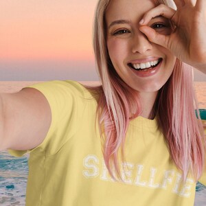 Puede incluir: Camiseta amarilla mostaza con la palabra "SWELLFIE" en letras blancas. La persona sonríe y se pone la mano delante de la cara. El fondo es una escena de playa con una puesta de sol.