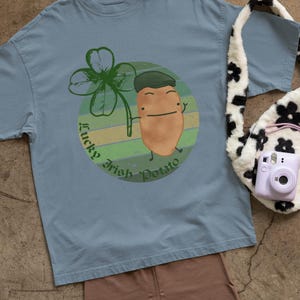 May include: A light blue t-shirt with a graphic of a potato wearing a green hat and a four-leaf clover. The shirt is paired with brown shorts, white sneakers, a gray beanie, and a black and white floral tote bag.