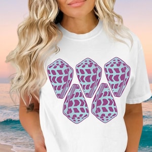 May include: A white short-sleeved t-shirt featuring a graphic print of five cone-shaped seashells arranged in a cluster. The shells are coloured in a light blue and purple pattern with a repeating geometric design across their surfaces.