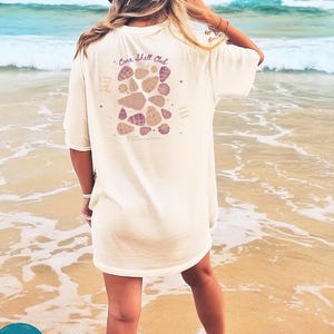 May include: Ivory t-shirt featuring a seashell graphic and the text "Cone Shell Club." The oversized shirt is worn on a beach. The text "some space open here" is also printed on the shirt. The sea is visible in the background.