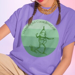 May include: A lilac t-shirt featuring the words "Shenanigan Coordinator" around a green circle with a cartoon character. The person is wearing beige trousers, white trainers, and a red watch.