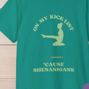 May include: A jade green t-shirt with a round neck. The shirt features a light green graphic with the text "ON MY KICK LIST" above a silhouette of a person doing a yoga pose, and the text "'CAUSE SHENANIGANS" below.