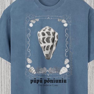 May include: Royal blue t-shirt with a seashell illustration inside a decorative frame. The shirt features the text "pūpū pōniuniu" and "The Hebrew Cone". The t-shirt is made of a soft material and is a relaxed fit.
