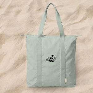 May include: A light sage green tote bag featuring a zipper closure at the top and two sturdy shoulder straps. The front of the bag displays a small, centered black embroidered graphic of a seashell with a distinct geometric pattern.
