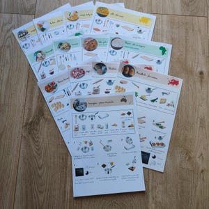 Könnte beinhalten: Ein Stapel bedruckter Rezeptkarten mit farbenfrohen Illustrationen und Text. Jede Karte zeigt ein anderes Gericht mit Schritt-für-Schritt-Anleitungen und Bildern von Zutaten und Kochutensilien. Die Karten sind auf einer Holzoberfläche angeordnet.