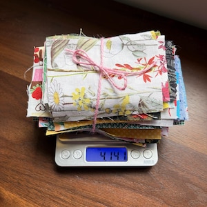 May include: A stack of fabric squares with floral patterns, tied with pink twine, rests on a digital scale. The scale displays a weight of 4.14 ounces. The fabrics feature various colors and designs, including flowers and leaves.