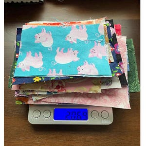 May include: A stack of fabric squares in various patterns, including a teal fabric with pink pig illustrations. The fabrics are stacked on a digital scale, displaying a weight of 206.9. Other patterns include floral and geometric designs.