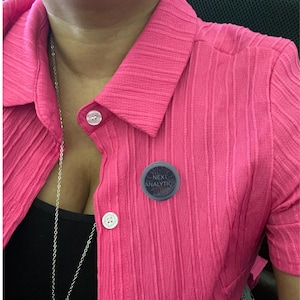 May include: A bright pink collared shirt with vertical ribbed texture and white buttons. A dark gray pin with the text "NEXT ANALYTICS" is pinned to the shirt. A silver necklace is visible.