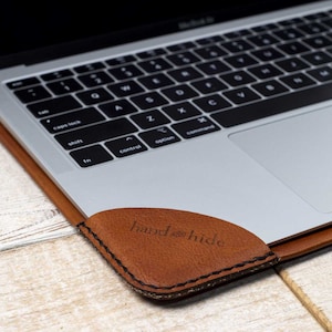 May include: A brown leather laptop sleeve with a black keyboard and a silver trackpad. The sleeve has the words "hand & hide" embossed on the corner. The laptop is open on a light wood surface.