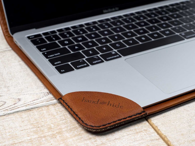 May include: A brown leather laptop sleeve with a black keyboard and a silver trackpad. The sleeve has the words "hand & hide" embossed on the corner. The laptop is open on a light wood surface.