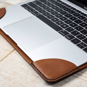 May include: A silver laptop with a black keyboard rests inside a brown leather sleeve. The sleeve has rounded corners and is stitched with black thread. The laptop is on a light wood surface.