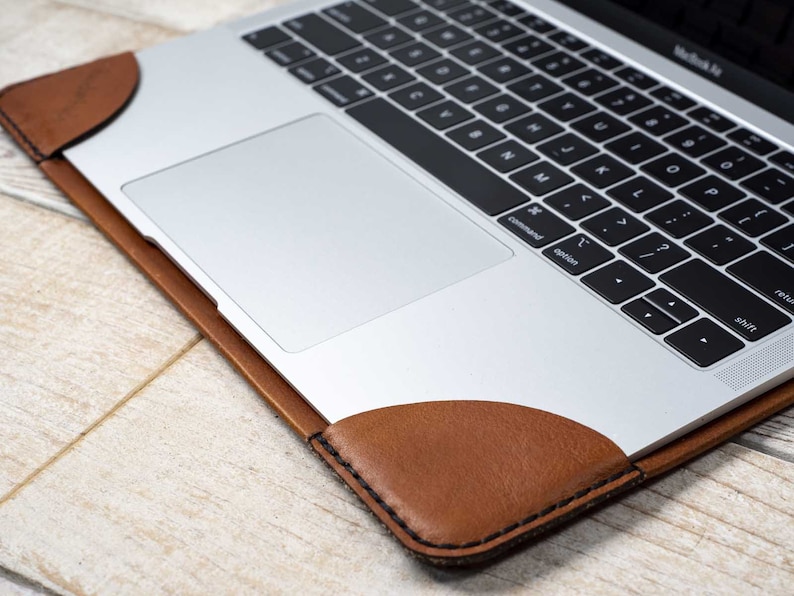 May include: A silver laptop with a black keyboard rests inside a brown leather sleeve. The sleeve has rounded corners and is stitched with black thread. The laptop is on a light wood surface.