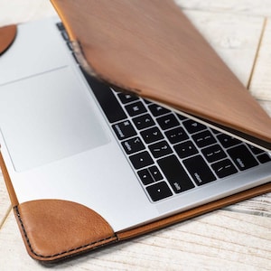 May include: A brown leather laptop sleeve with a laptop inside. The sleeve has rounded corners and a stitched border. The laptop is silver with a black keyboard. The sleeve is open, revealing the laptop's keyboard and trackpad.